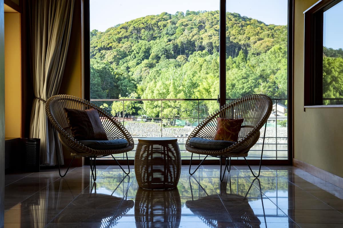 A room with two wicker chairs and a small table, large windows with a view of a forested hill.