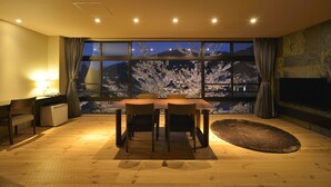 A dining area with a table and chairs, a small refrigerator, a lamp, and a view of cherry blossoms through large windows.