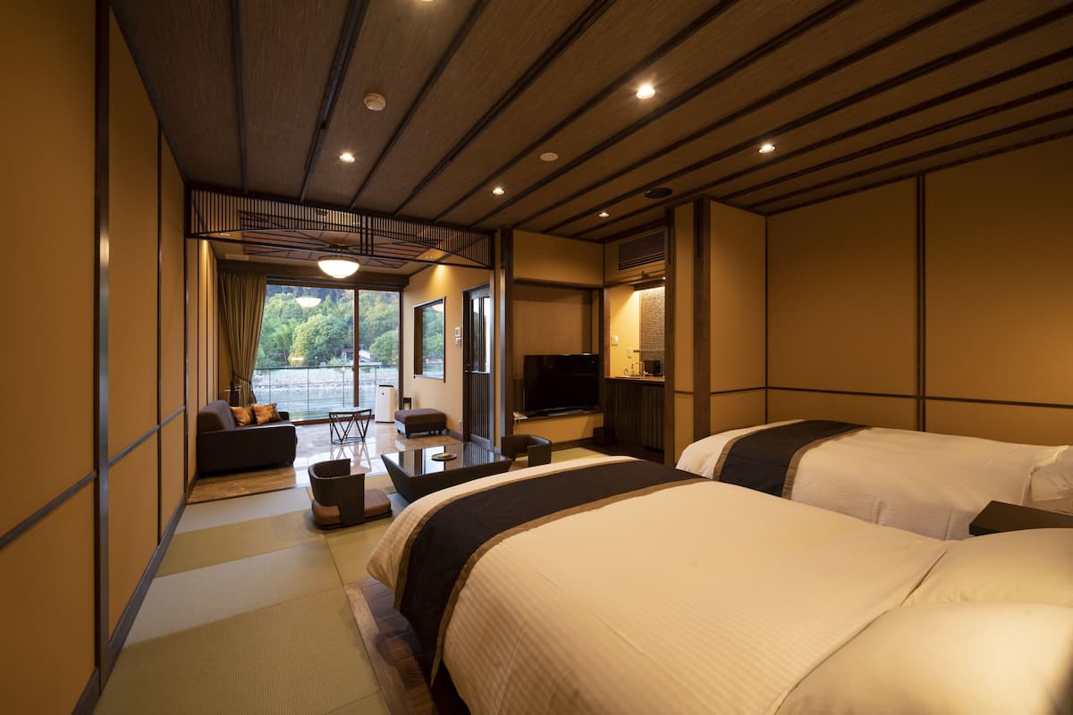 A hotel room with two beds, a sofa, a TV, and a view of greenery outside.