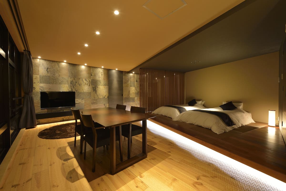 A modern hotel room with a wooden dining table, a flat-screen TV, a sofa bed, and a stone-patterned wall.