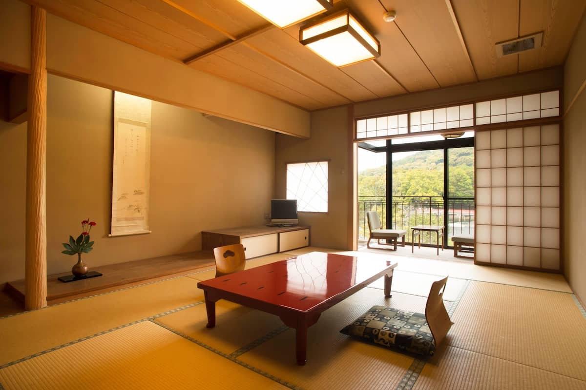 A traditional Japanese room with tatami flooring, a low wooden table, a television on a cabinet, and a sliding door leading to a balcony with a view of greenery.