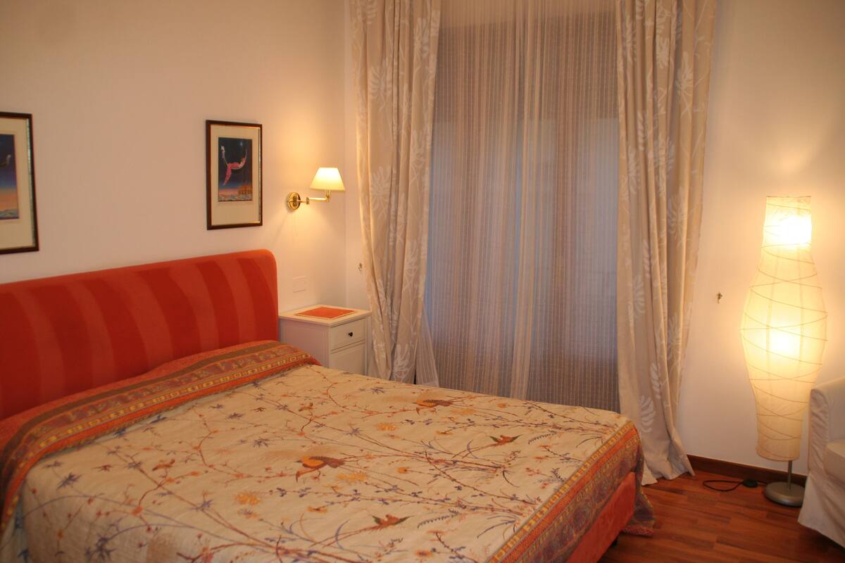 Quarto com cama, mesa de cabeceira, luminária e janela com cortinas.