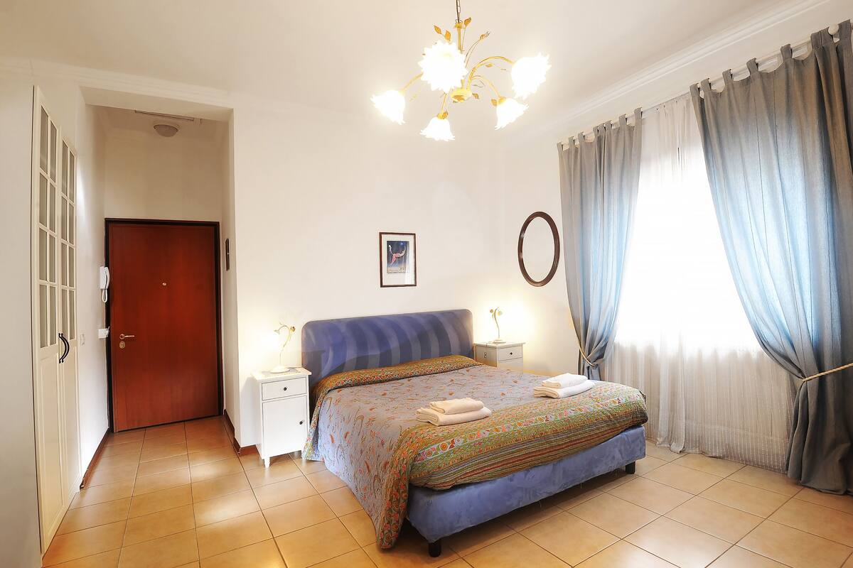 Quarto com cama, mesas de cabeceira, um candelabro, uma porta e cortinas.