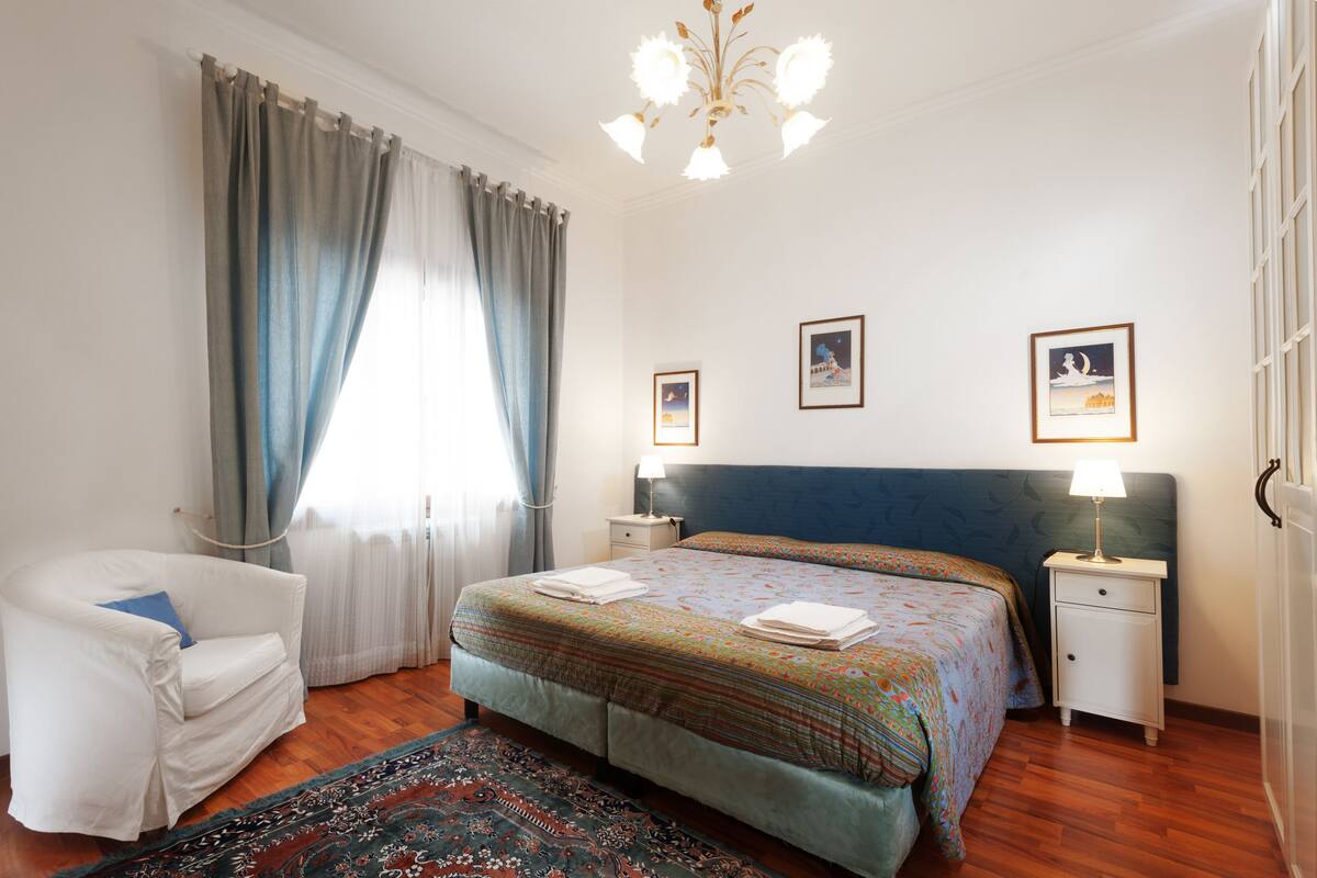 Quarto com uma cama grande, uma cadeira, uma mesinha de cabeceira e um candelabro.