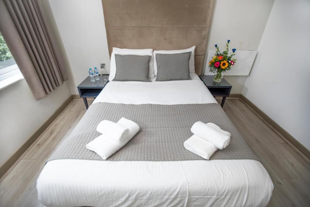 Een netjes opgemaakt bed met twee kussens, een nachtkastje met waterflesjes en een vaas met bloemen.