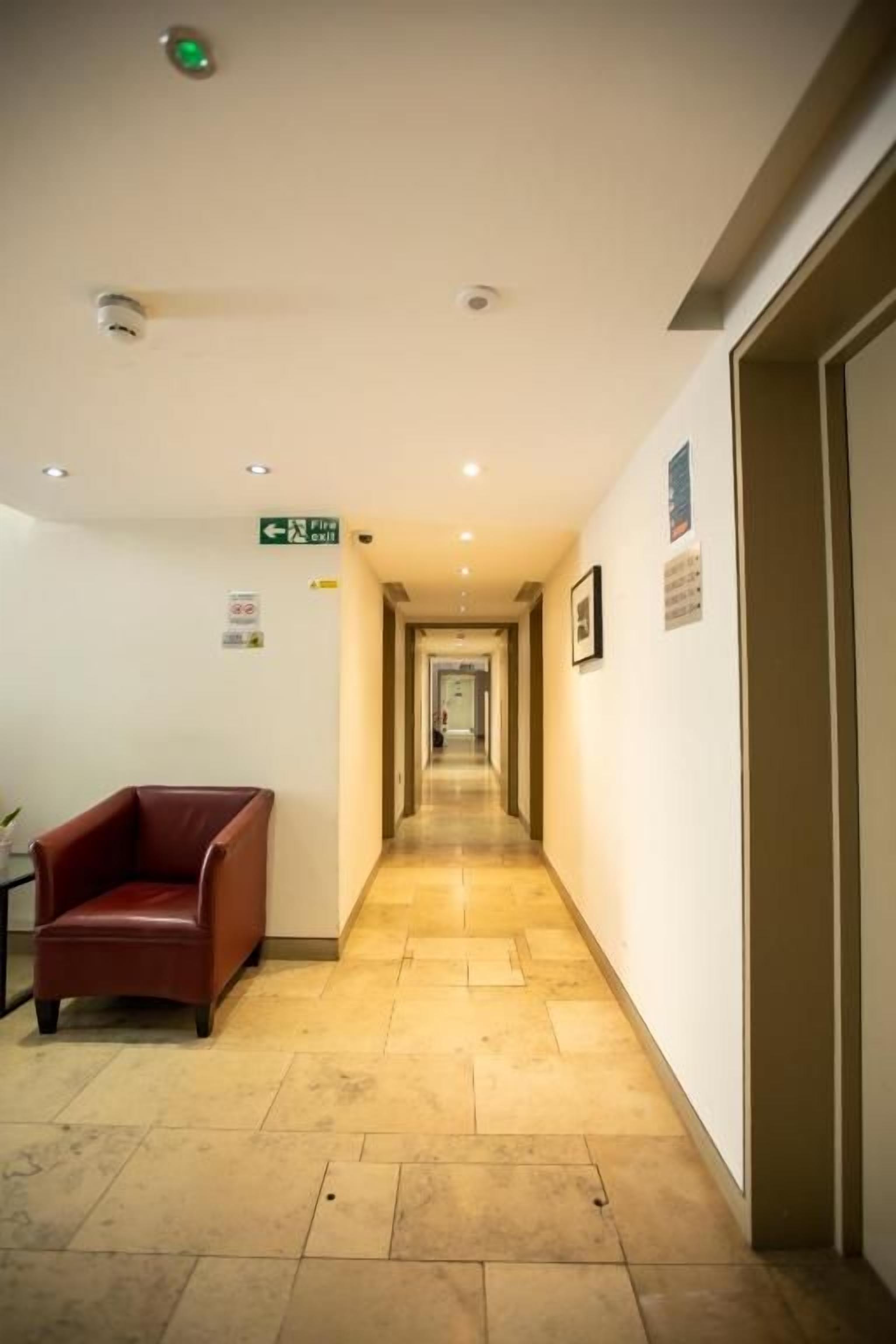 A hotel corridor with a red armchair, a fire exit sign, and a framed picture on the wall.