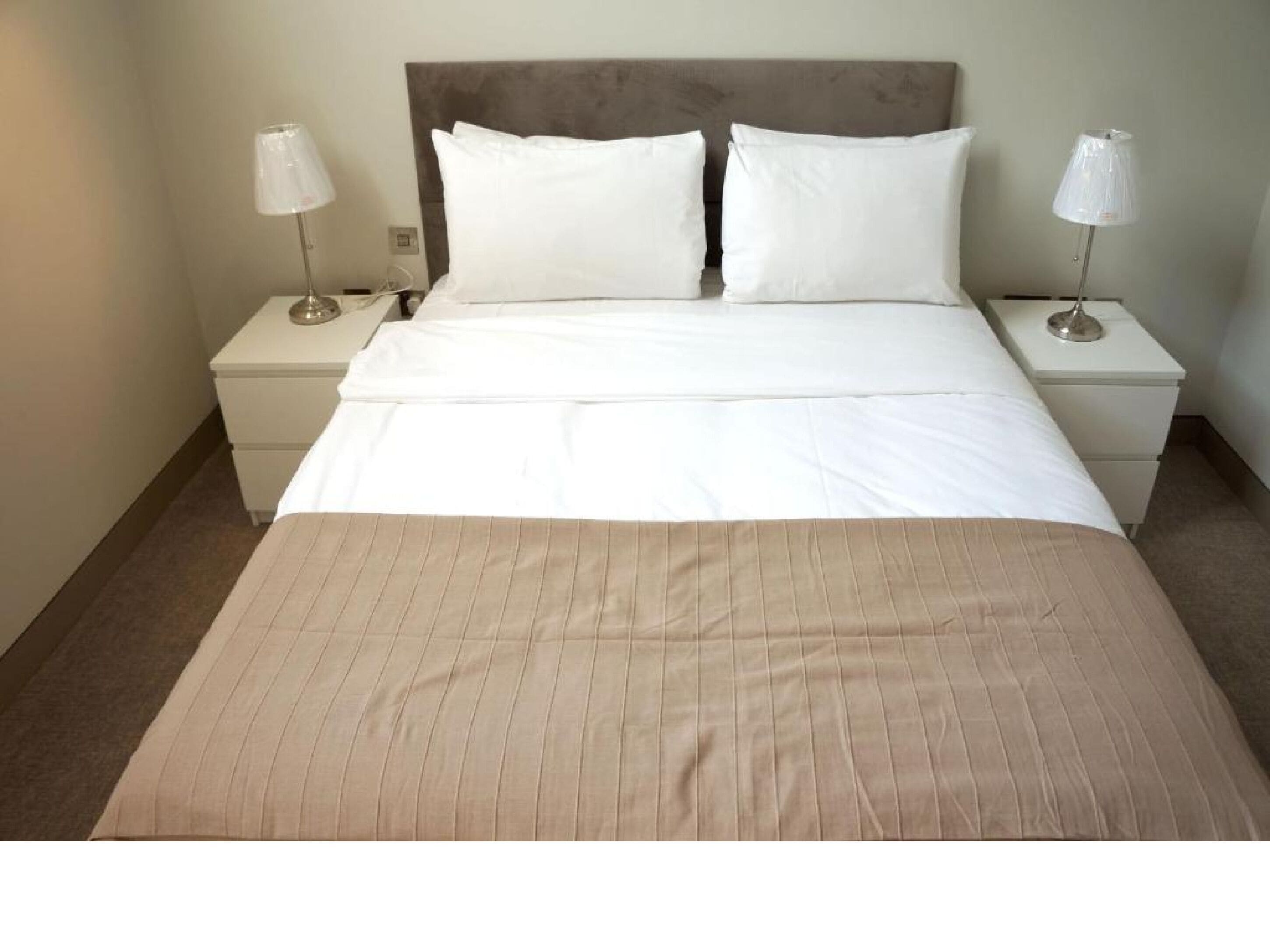 A neatly made bed with white linens and a beige bedspread, flanked by two bedside tables with lamps.