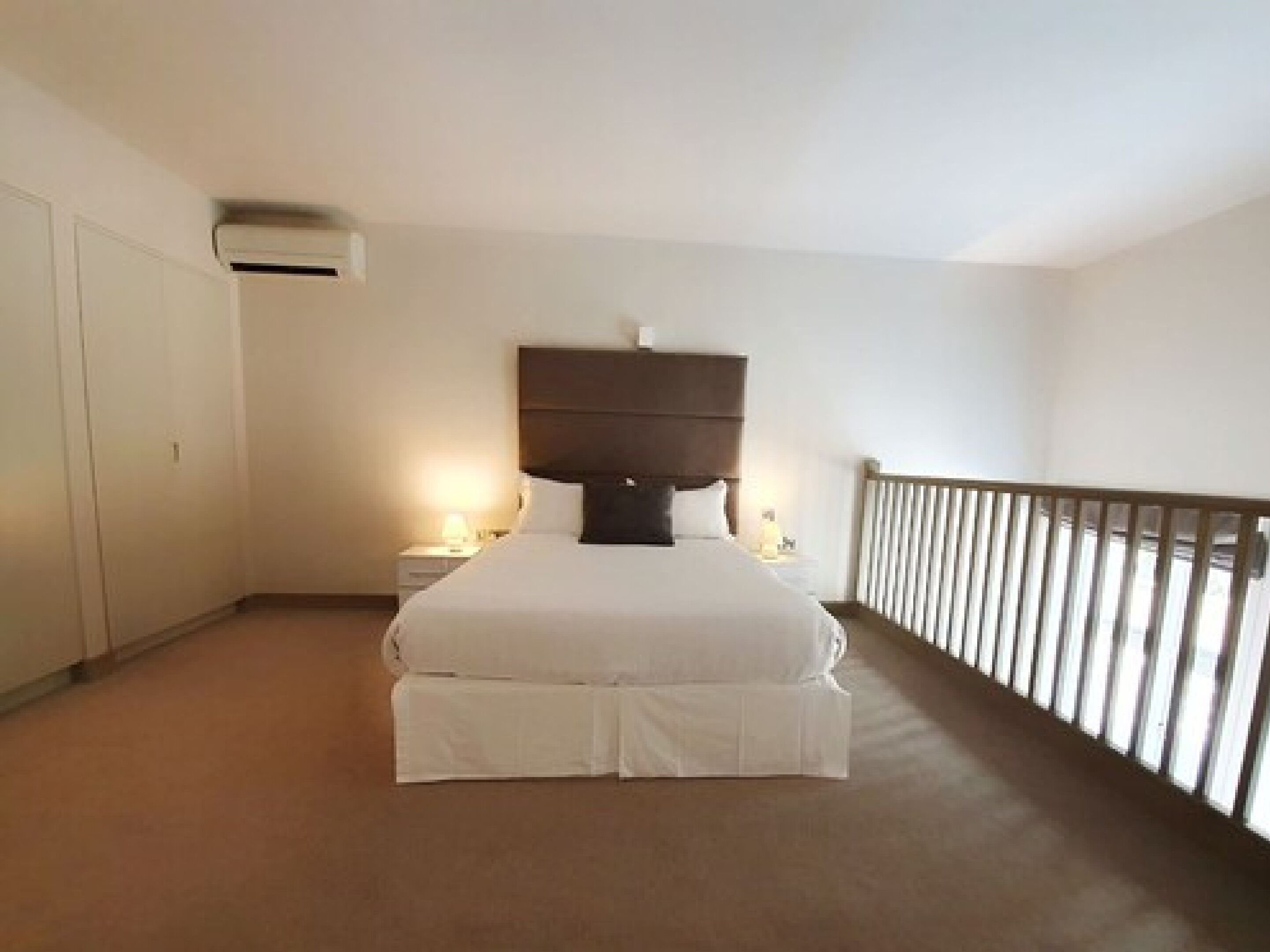 Una habitación de hotel con una cama grande, una mesita de noche, aire acondicionado montado en la pared y un balcón con barandilla.