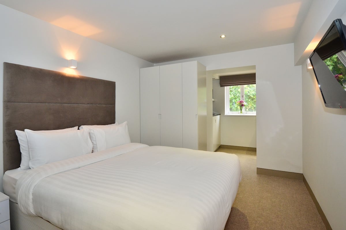 Ein modernes Hotelzimmer mit einem großen Bett, einem Kopfteil, einem an der Wand befestigten Flachbildfernseher und einem Fenster mit Blick auf Grünflächen.