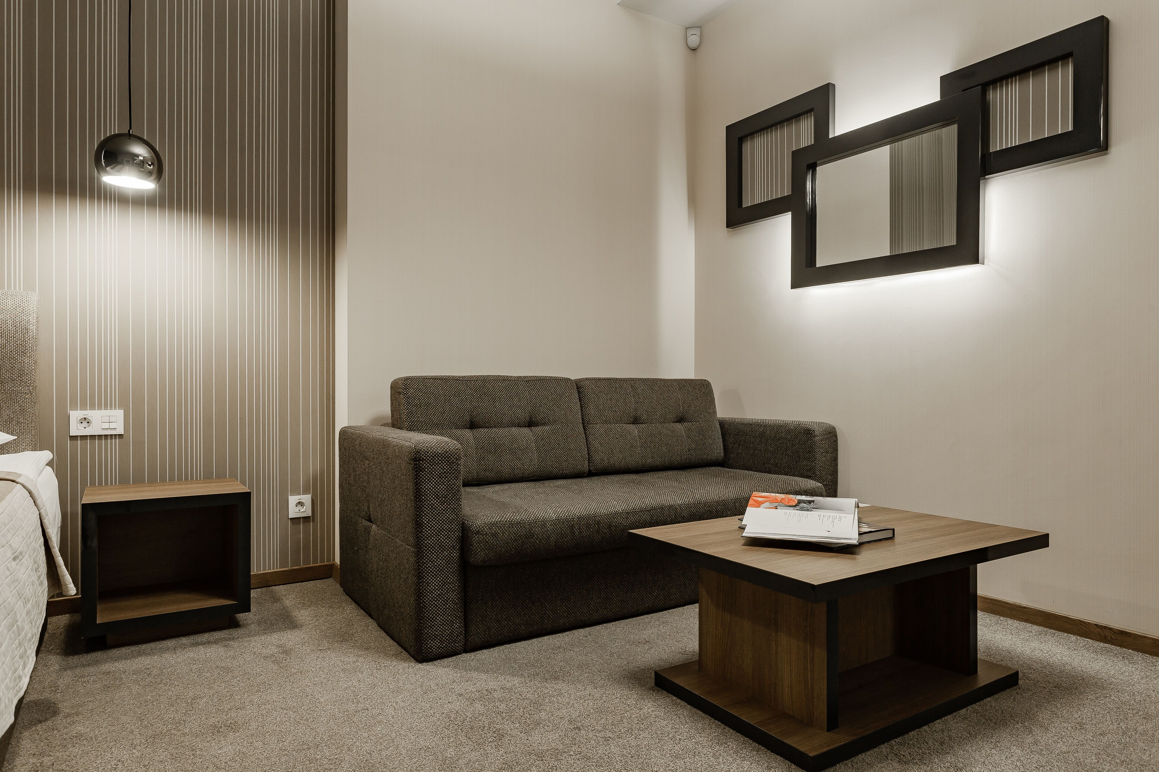 A modern hotel room with a brown sofa, a wooden coffee table, a small side table, and a wall-mounted mirror.