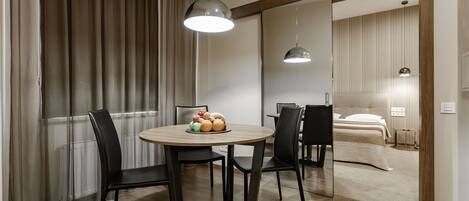 Una habitación de hotel moderna con un área de comedor que incluye una mesa redonda, sillas y un frutero. La habitación tiene piso de madera, una cama con cabecera blanca y un ventanal con cortinas.