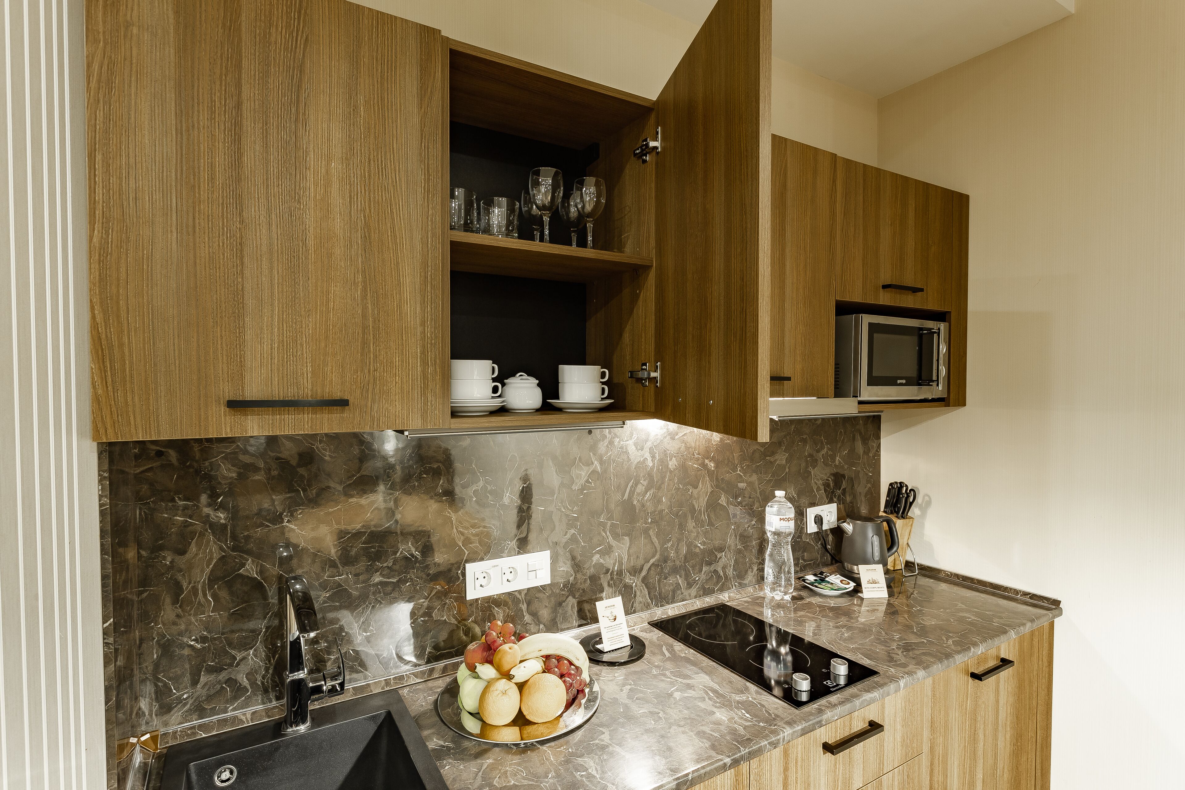 A modern kitchen with wooden cabinets, a marble backsplash, a sink, a microwave, and a countertop with a fruit bowl.