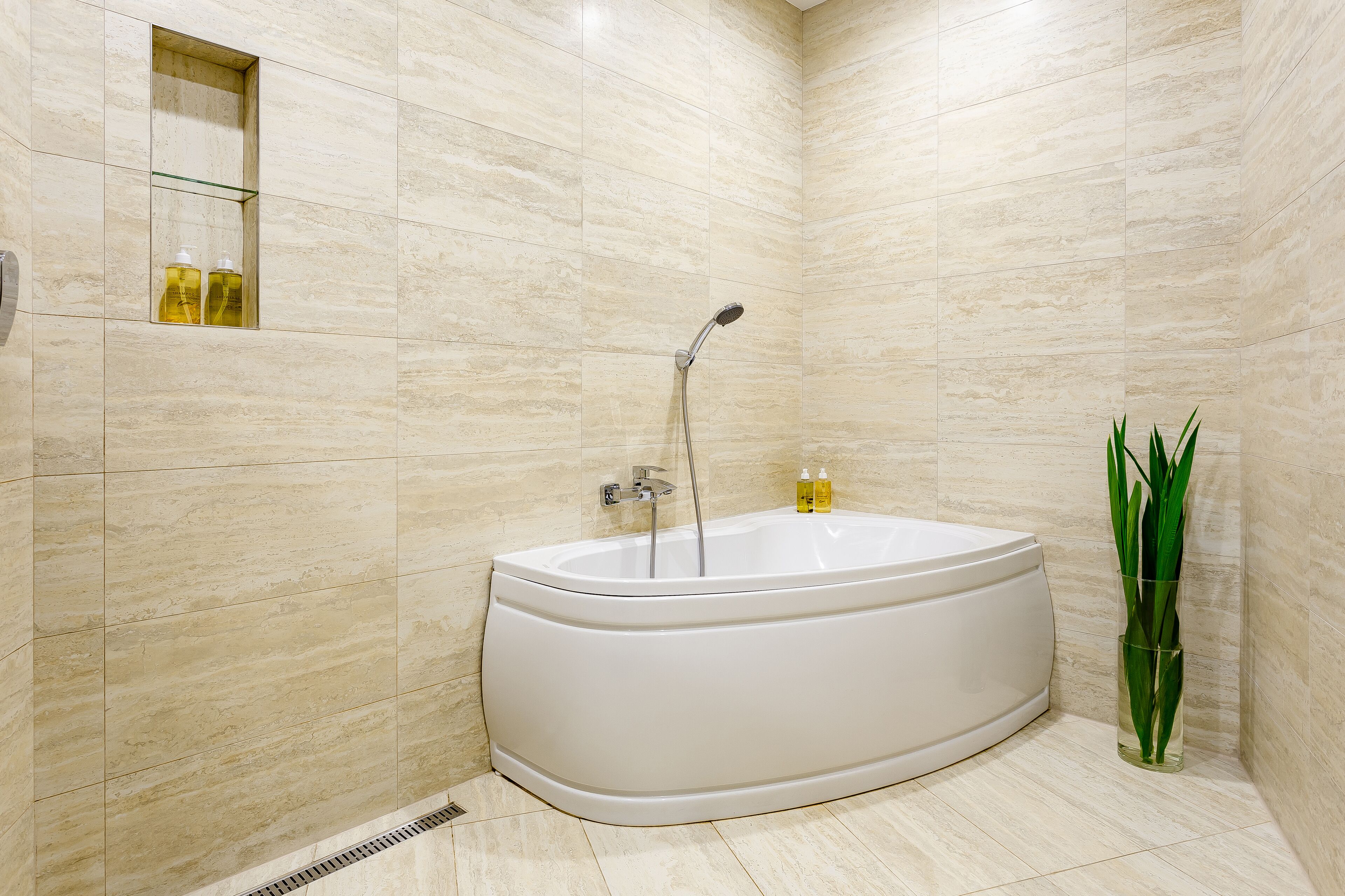 A modern bathroom with a freestanding bathtub, a glass shelf with toiletries, and a tall green plant.
