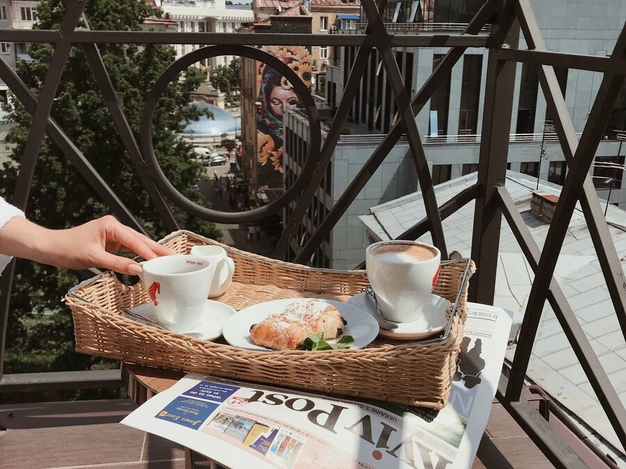 A wicker tray with two cups of coffee, a pastry, and a newspaper on a balcony railing.