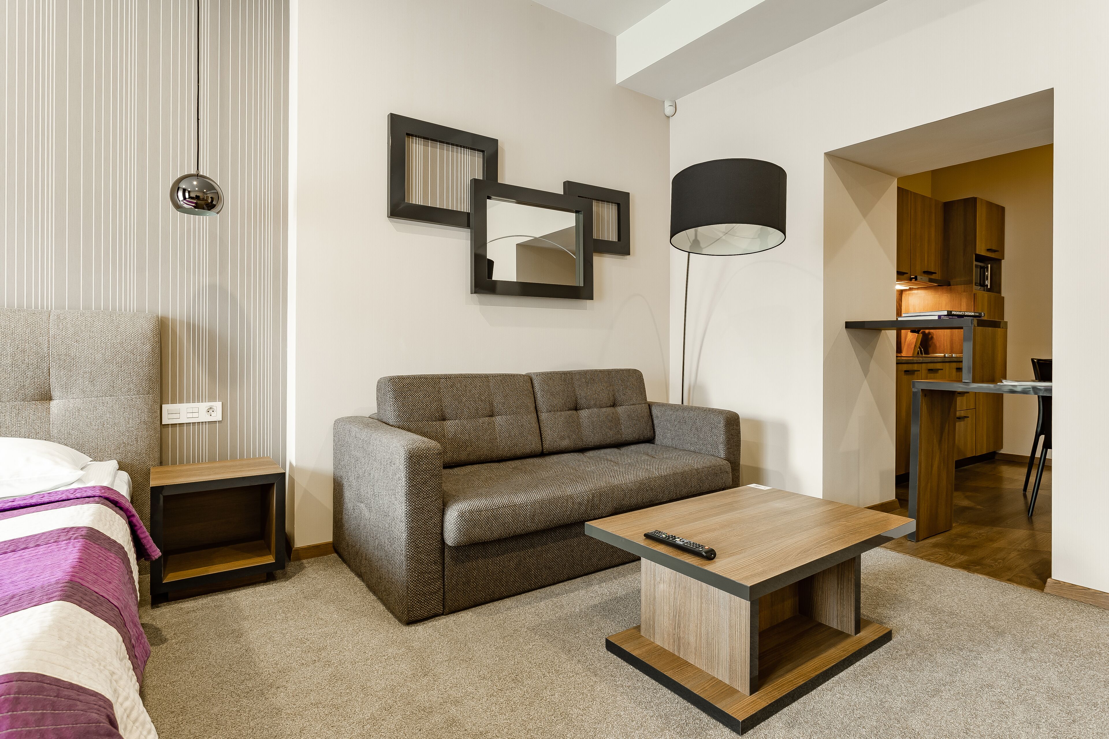 A modern hotel room with a sofa, a small table, a bedside table, and a lamp.