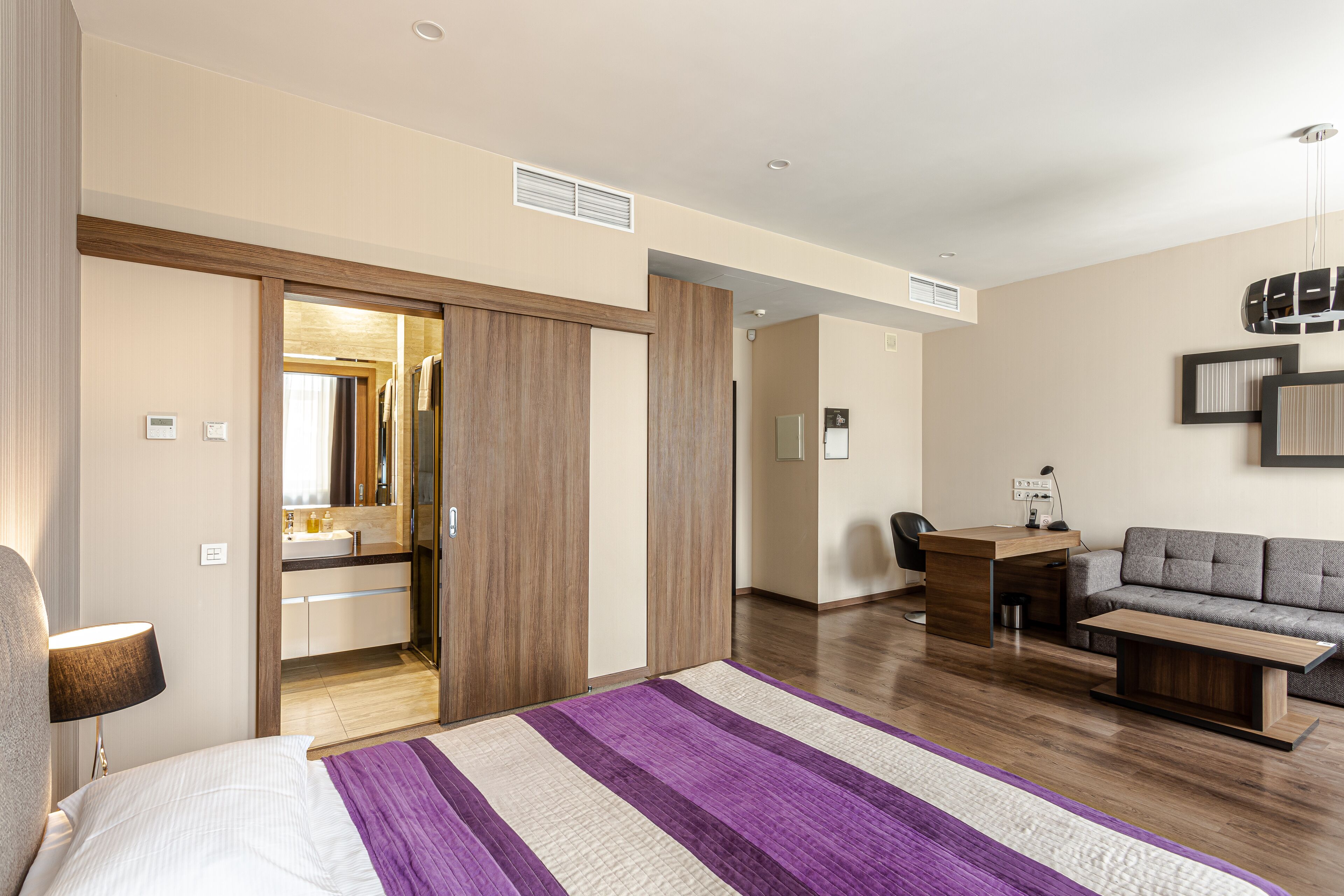 A modern hotel room with a bed, a desk, a sofa, and a bathroom visible through an open door.