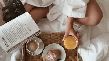 Una persona con bata blanca sentada en una cama con una bandeja que contiene un croissant, una taza de café y un vaso de jugo de naranja, sosteniendo un libro.