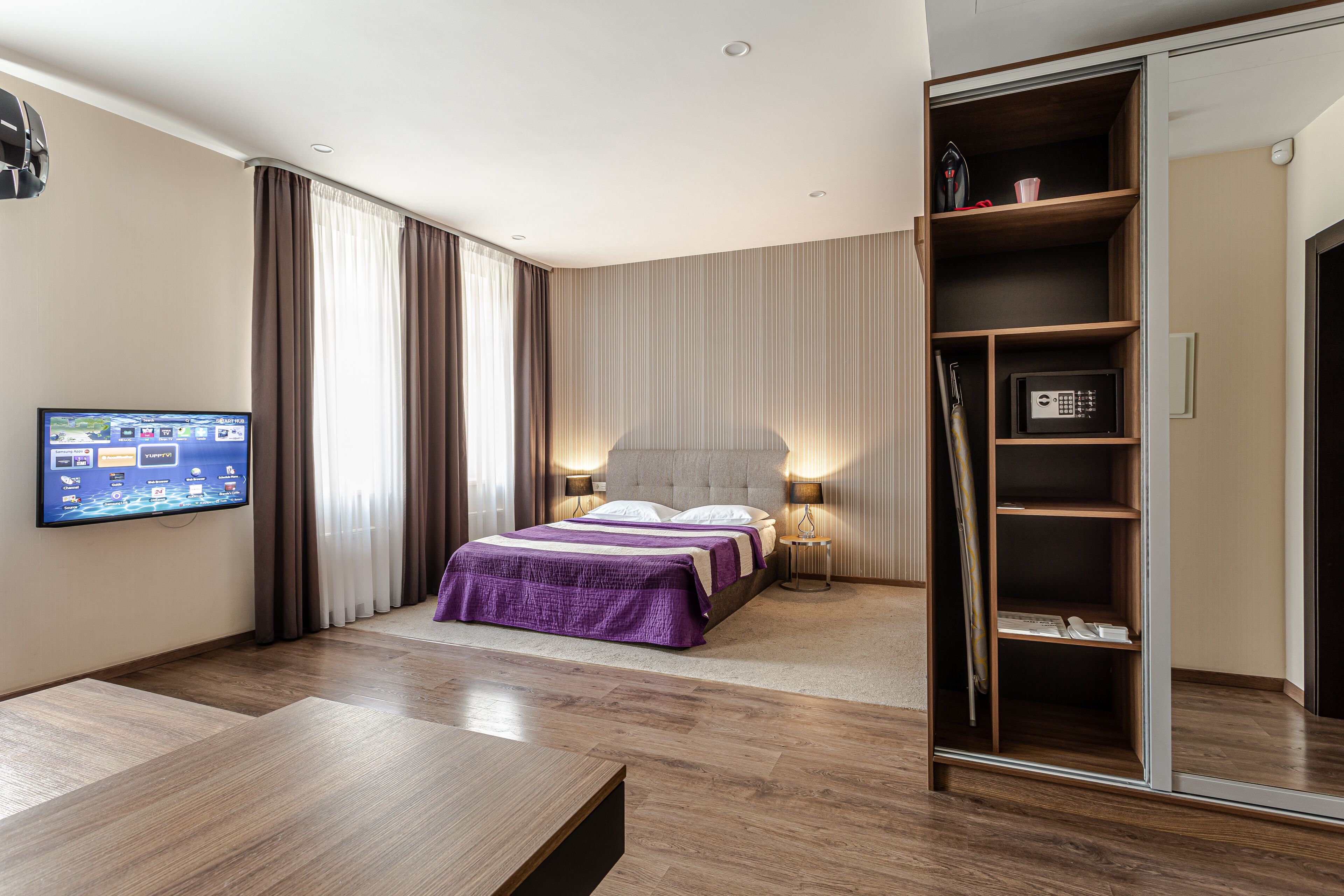 A modern hotel room with a large bed, a television mounted on the wall, a wardrobe, and a bedside table.