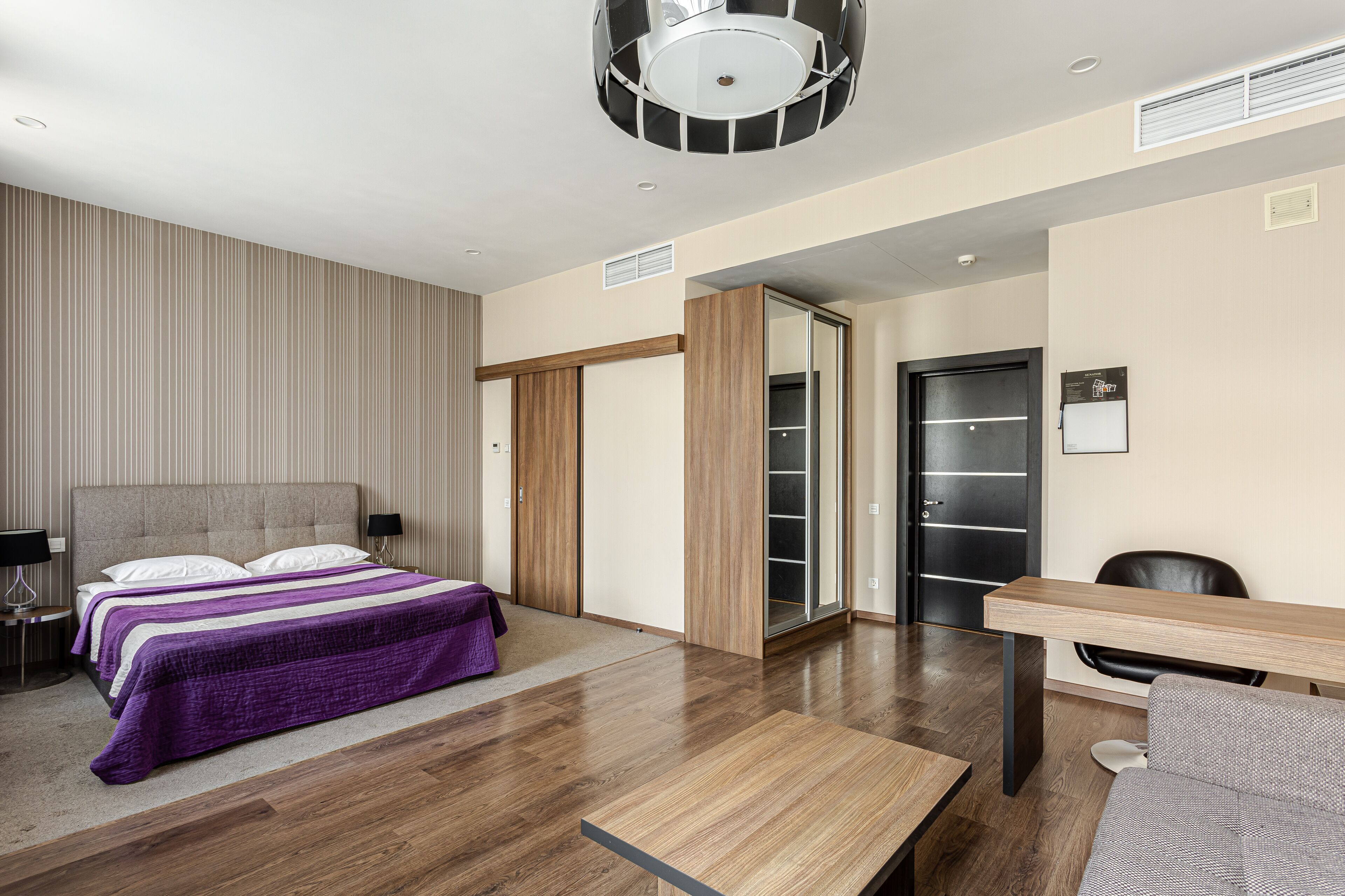 A modern hotel room with a large bed, a wooden desk, and a wardrobe.