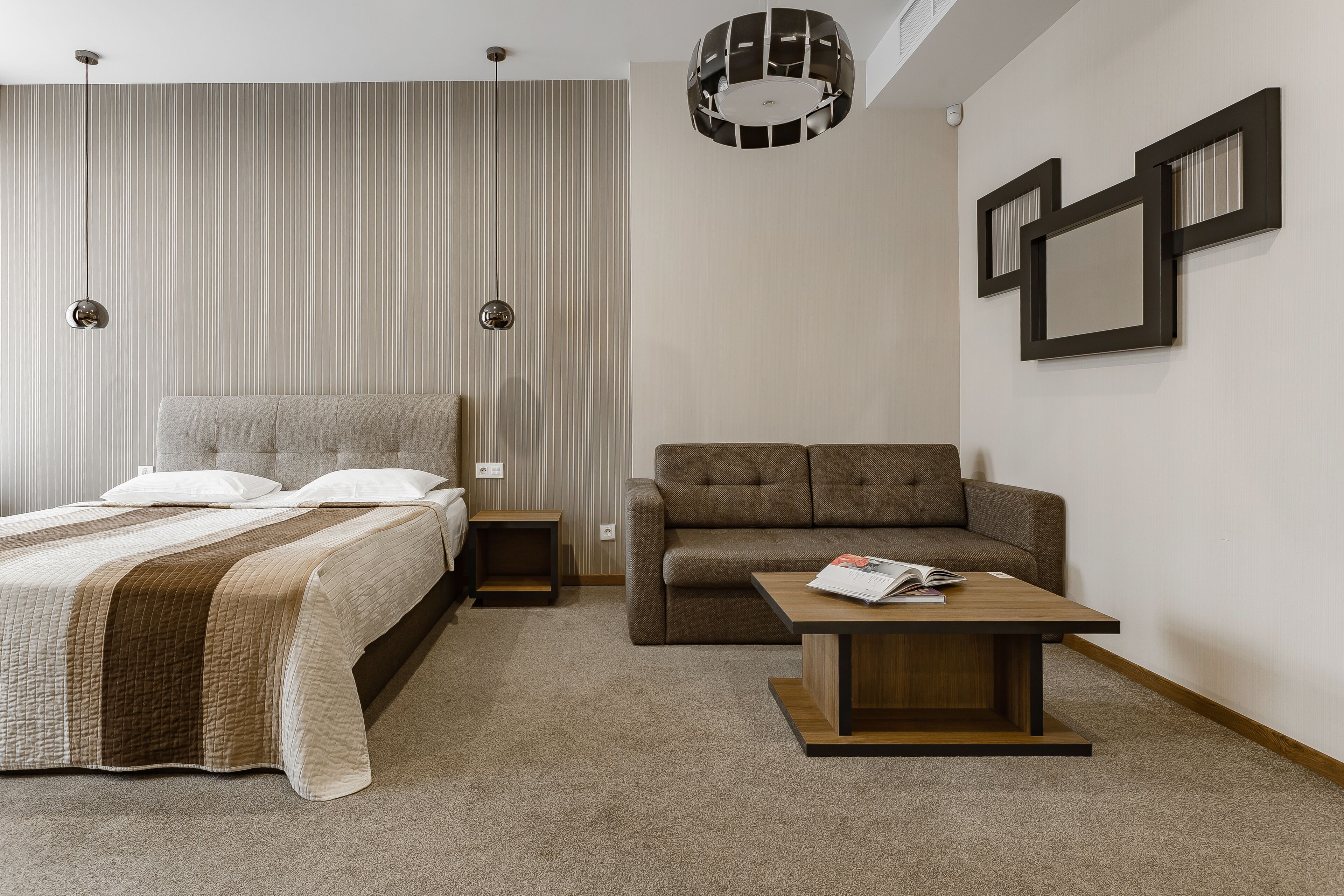A modern hotel room with a bed, a sofa, a coffee table, and a wall-mounted light fixture.