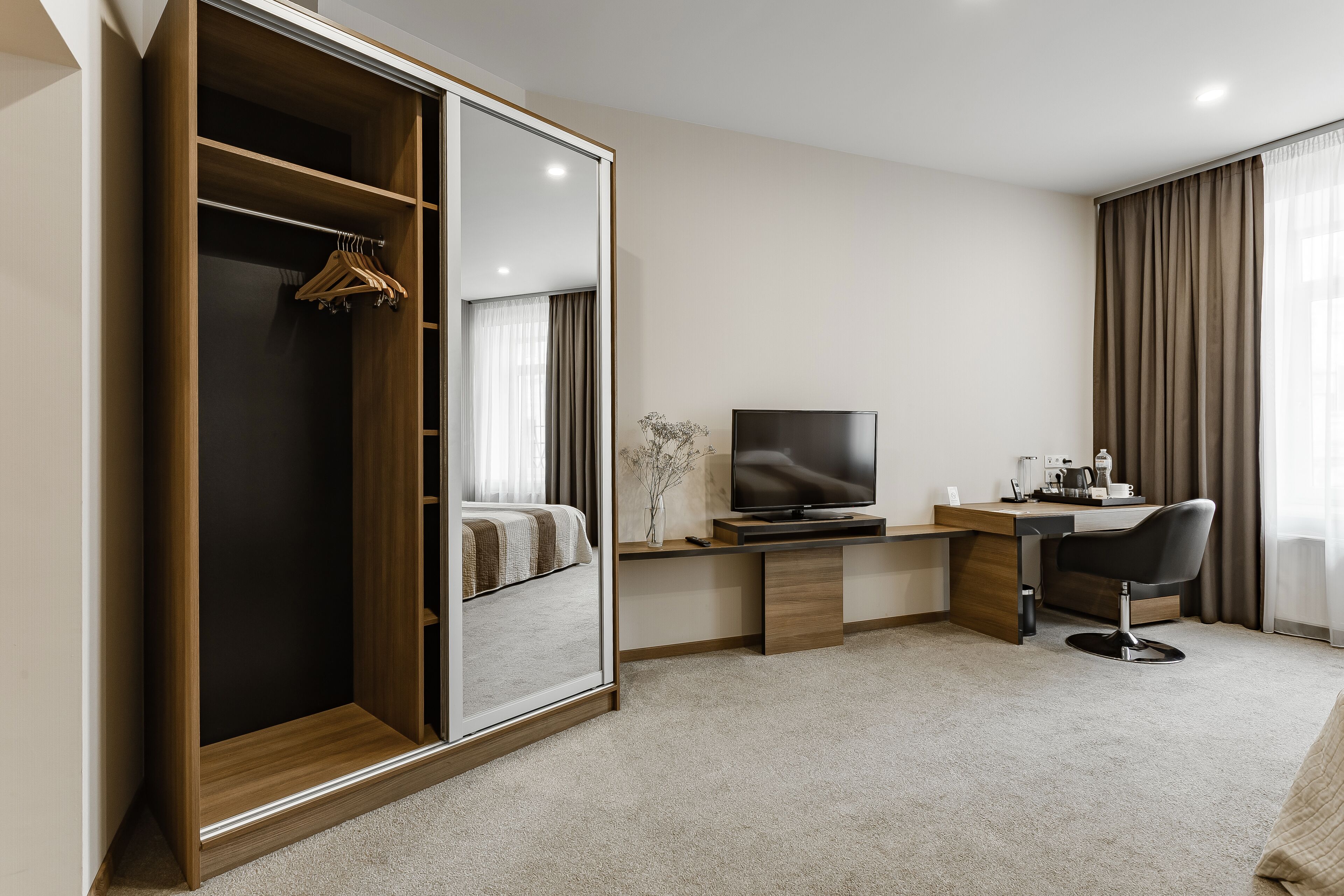 A modern hotel room with a walk-in closet, a flat-screen TV, a desk with a chair, and a large window with curtains.