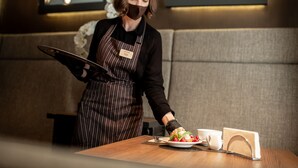 A person in a striped apron standing at a wooden table with a plate of food, a cup, and a menu stand. - Senator Maidan (Kyiv)