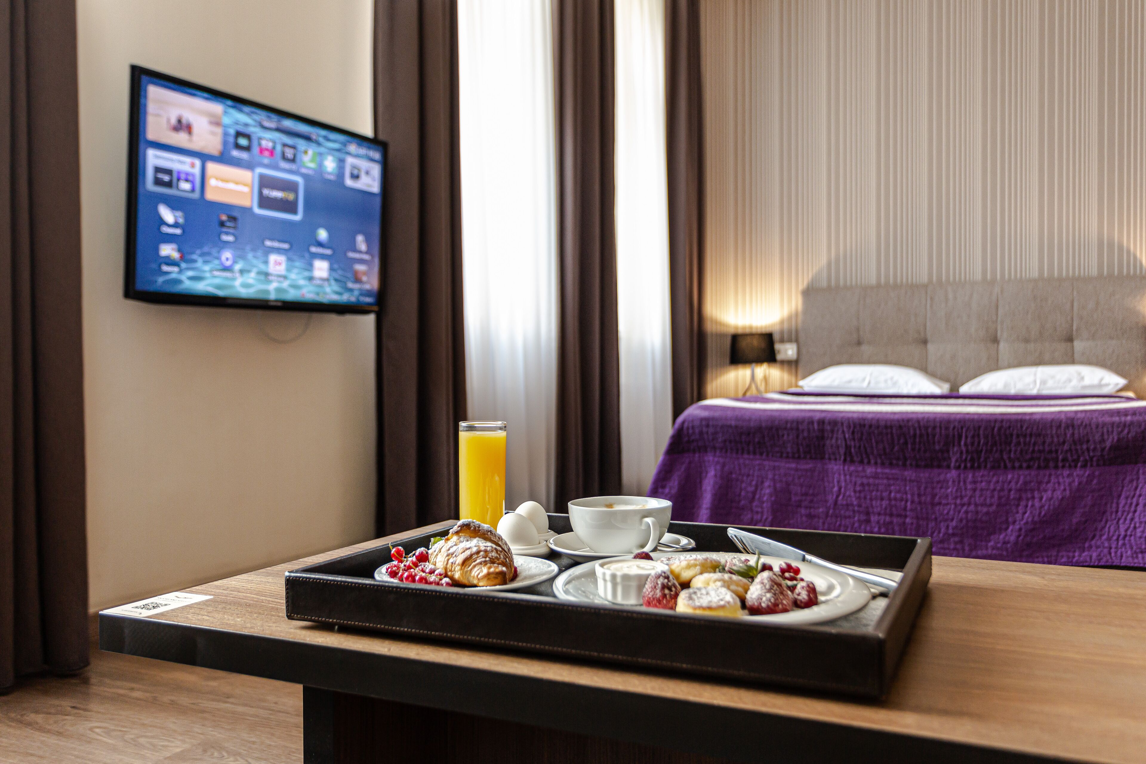 Een hotelkamer met een bed, een televisie aan de muur en een ontbijtkarretje met gebak en sap.