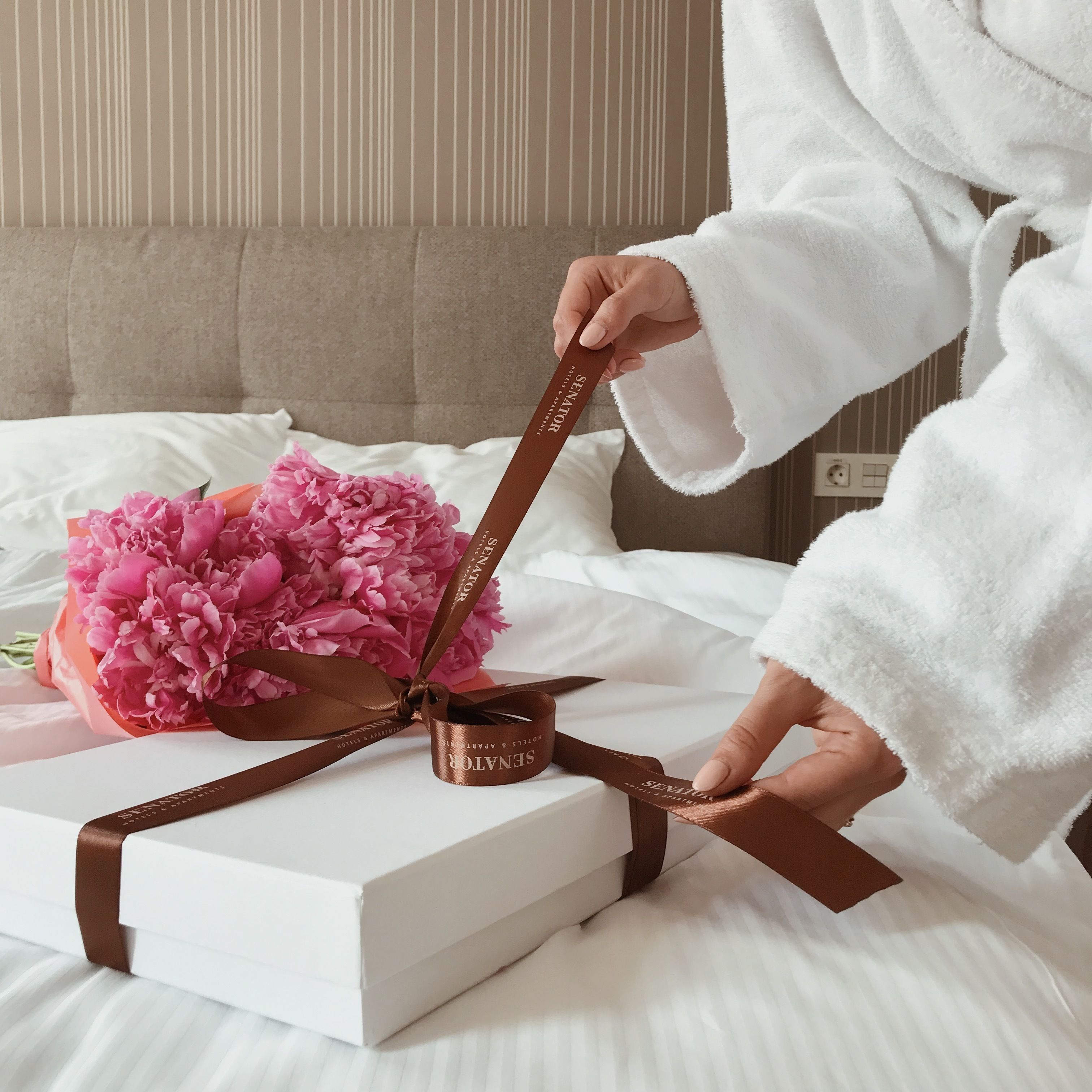 Une personne vêtue d’une robe blanche tient un ruban marron noué autour d’une boîte blanche, et à côté se trouve un bouquet de fleurs roses.