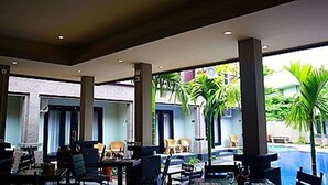 A covered outdoor seating area with tables and chairs, large pillars, and a view of a swimming pool and greenery. - Taman Tirta Ayu (Tuban)