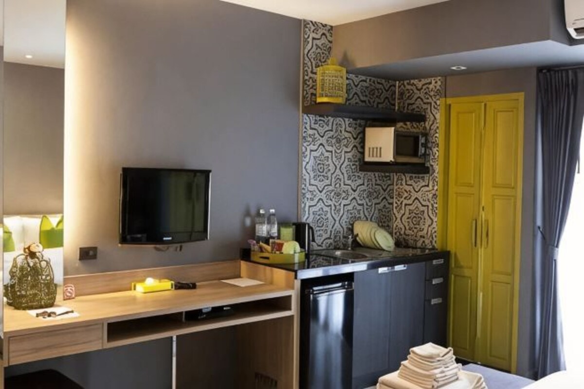 A compact kitchen with a microwave, a sink, and a small refrigerator. There is a television mounted on the wall, and a yellow door is visible in the background.