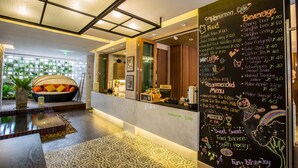 Un café avec un tableau noir affichant le menu des plats et des boissons, un espace salon avec un canapé et une plante, et un plafond décoratif.
