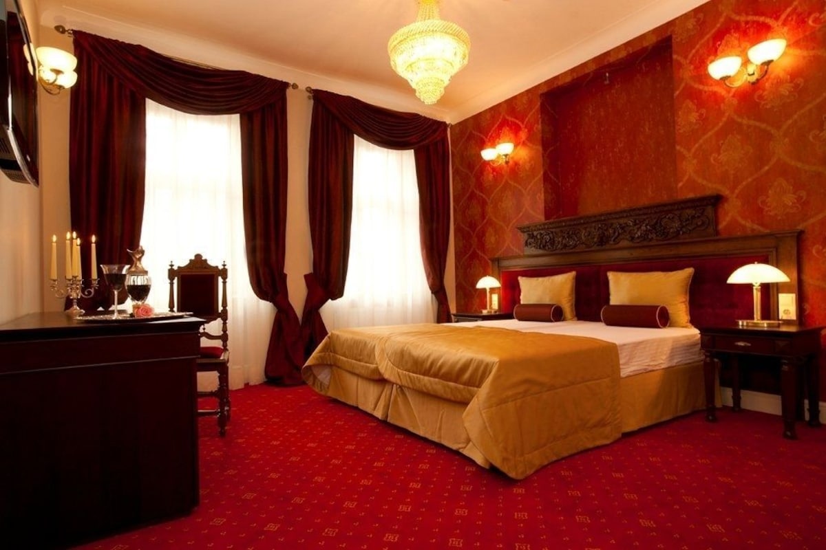 A hotel room with a large bed, a desk, a chandelier, and a red carpeted floor.