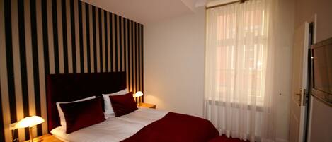 A hotel room with a bed, a nightstand, a lamp, a window with sheer curtains, and a door.