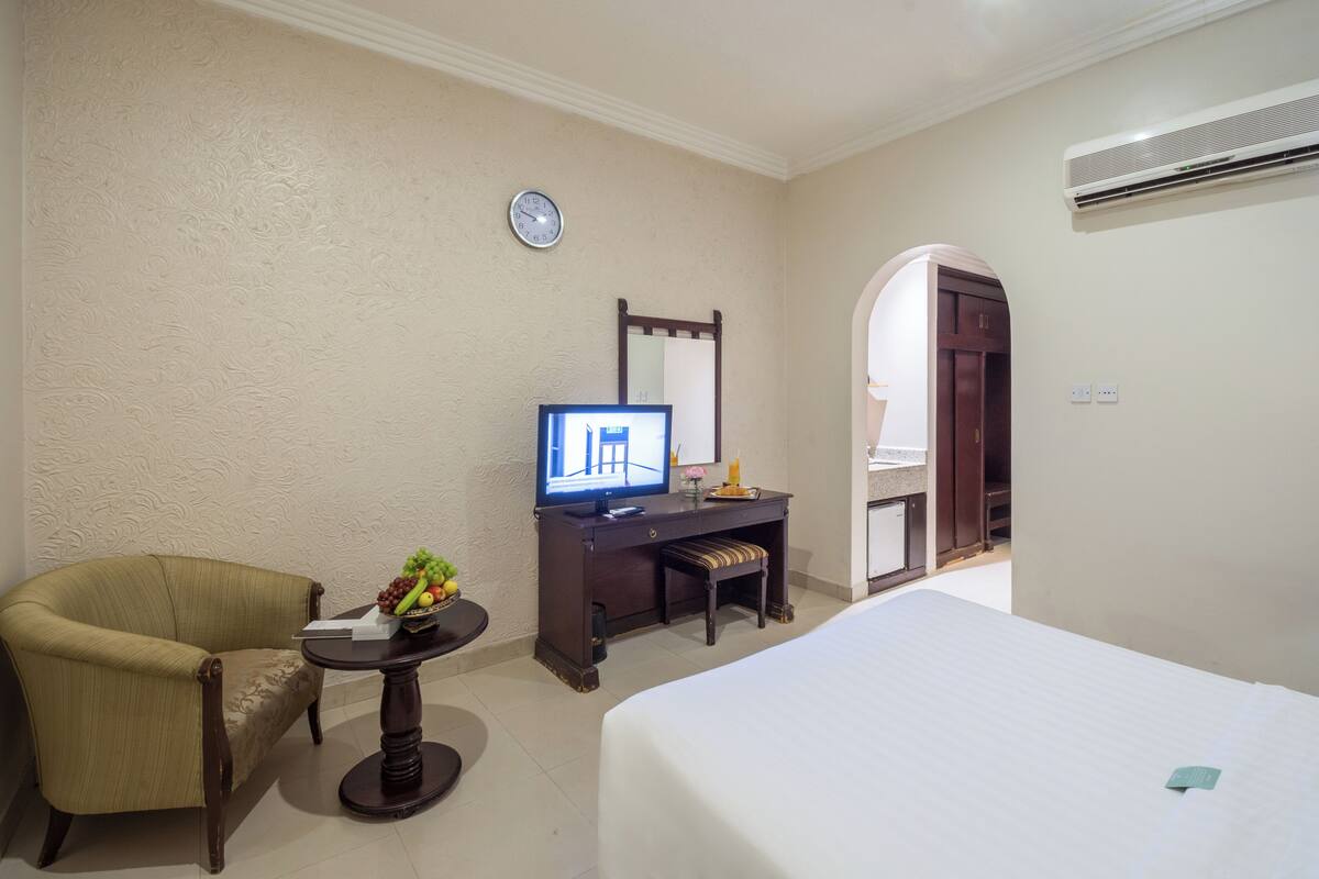 A hotel room with a bed, a desk with a computer, a chair, a clock, and an air conditioning unit.