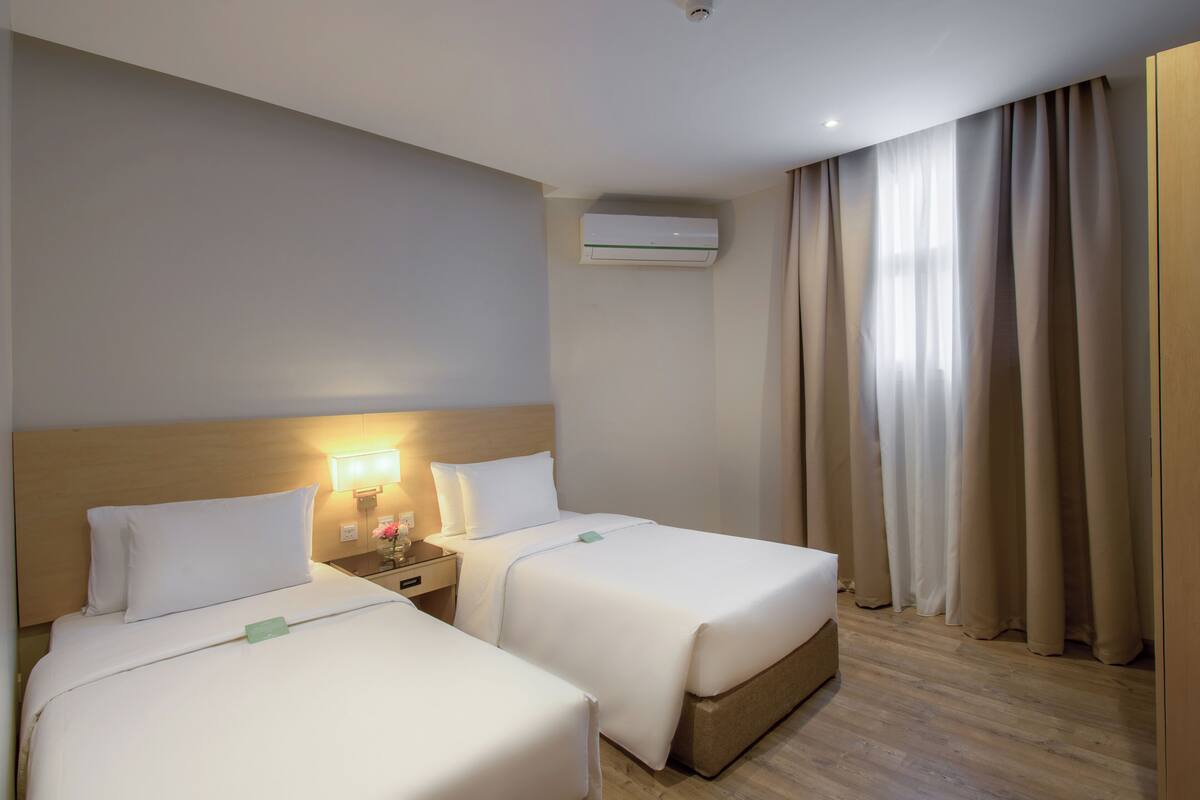A hotel room with two beds, a nightstand with a lamp, an air conditioning unit, and a window with curtains.