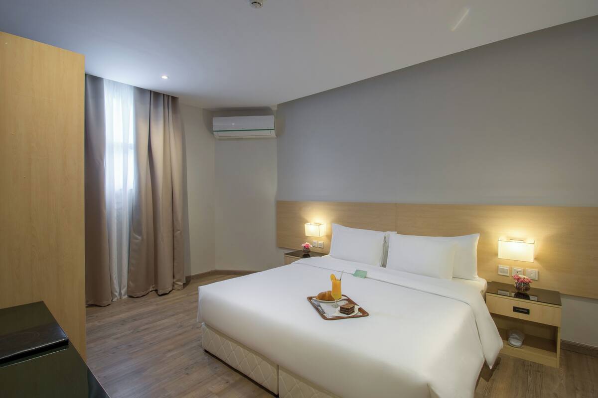 A hotel room with a large bed, bedside lamps, a nightstand, a small table with a tray, and a window with curtains.