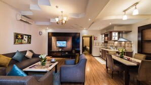 A modern living room with a sofa, armchairs, and a coffee table. There is a TV mounted on the wall and a kitchen area with a dining table and chairs. - Boudl Al Masif (Riyadh)