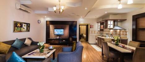 A modern living room with a sofa, armchairs, and a coffee table. There is a TV mounted on the wall and a kitchen area with a dining table and chairs.