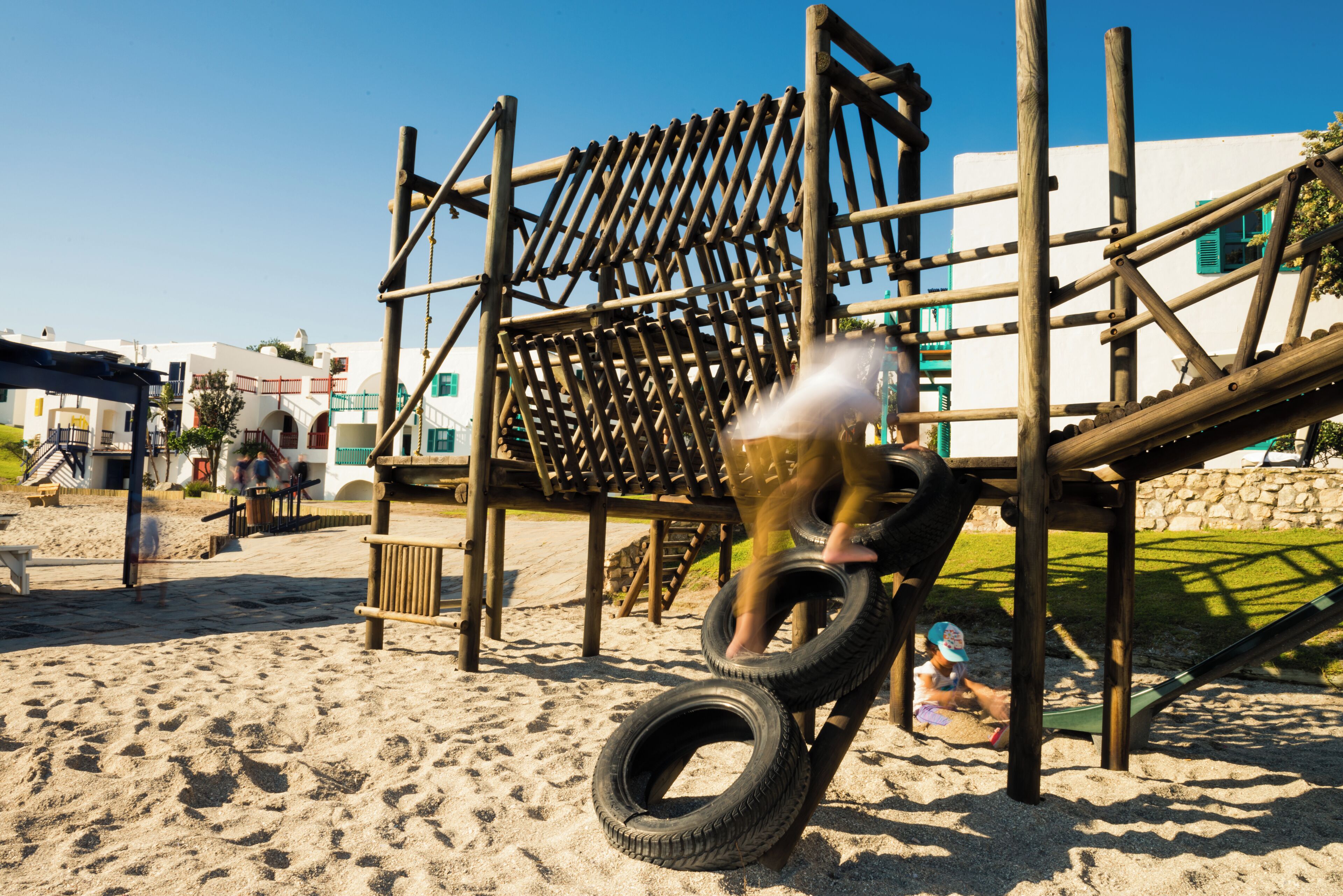 A wooden play structure with a slide and climbing elements, situated on a sandy beach with buildings in the background.
