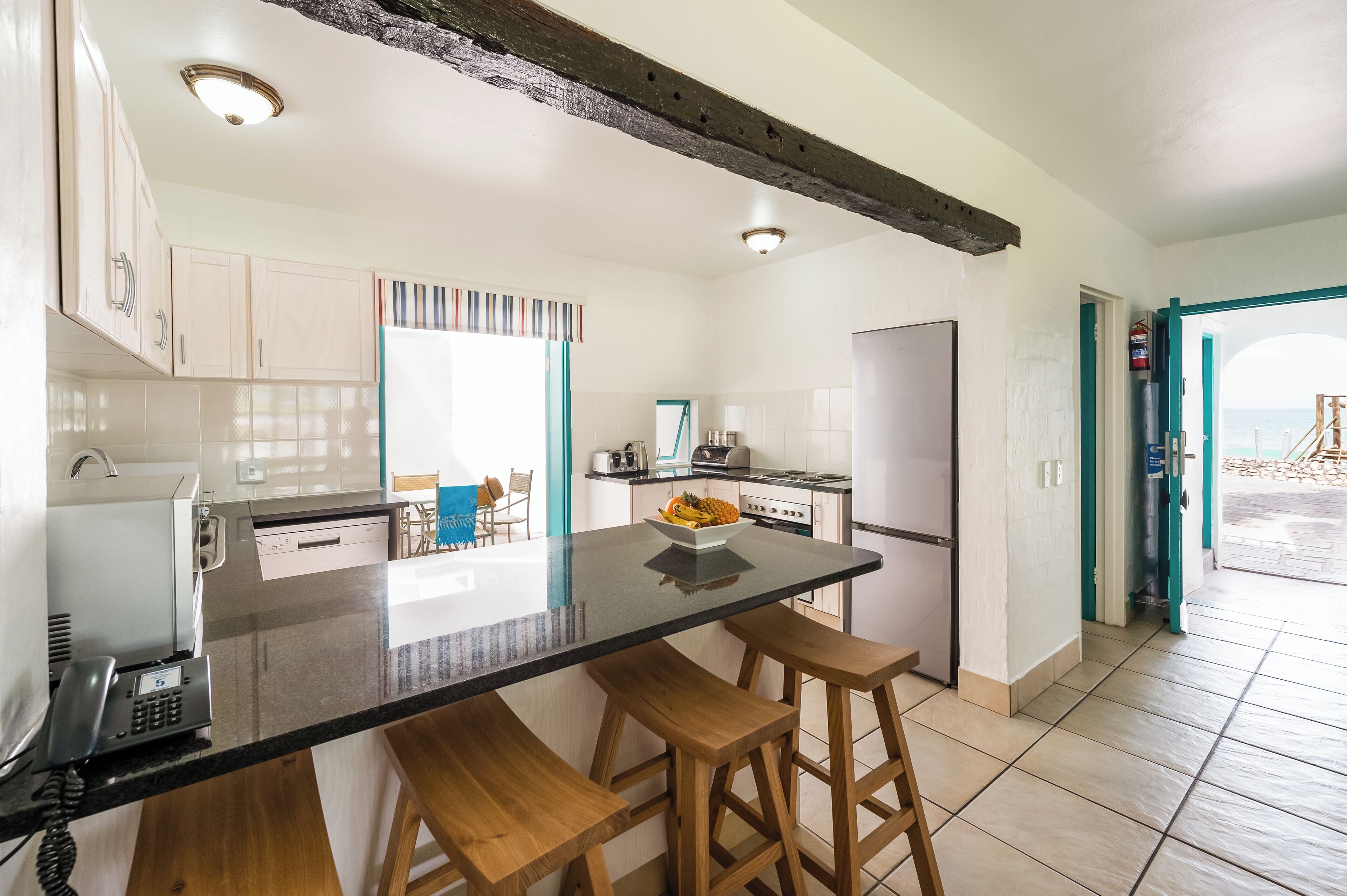 A modern kitchen with a central island, wooden stools, and a view of the sea through an open door.