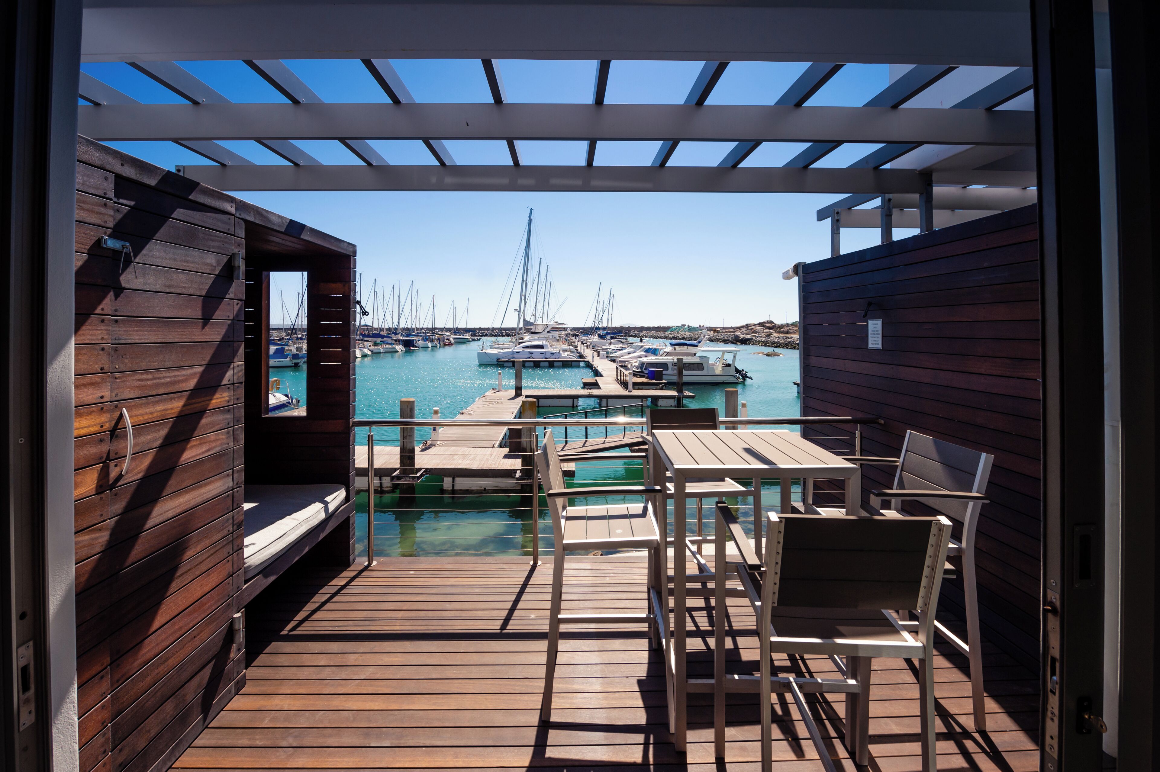 Una terraza de madera con una pérgola, con vistas a un puerto deportivo lleno de barcos y un cielo despejado.