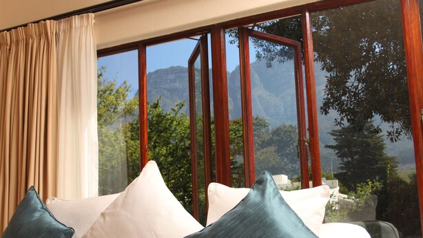 A room with a large window, a sofa with cushions, and a view of mountains. - Riversong Guest House (Cape Town)