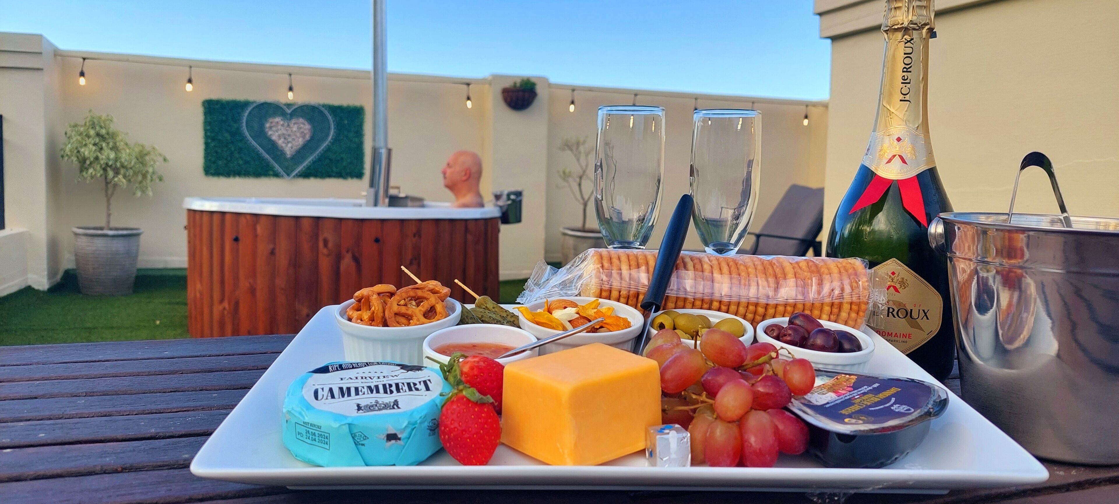 A picnic setup with a variety of cheeses, fruits, and snacks on a tray, a bottle of champagne, and a hot tub in the background.