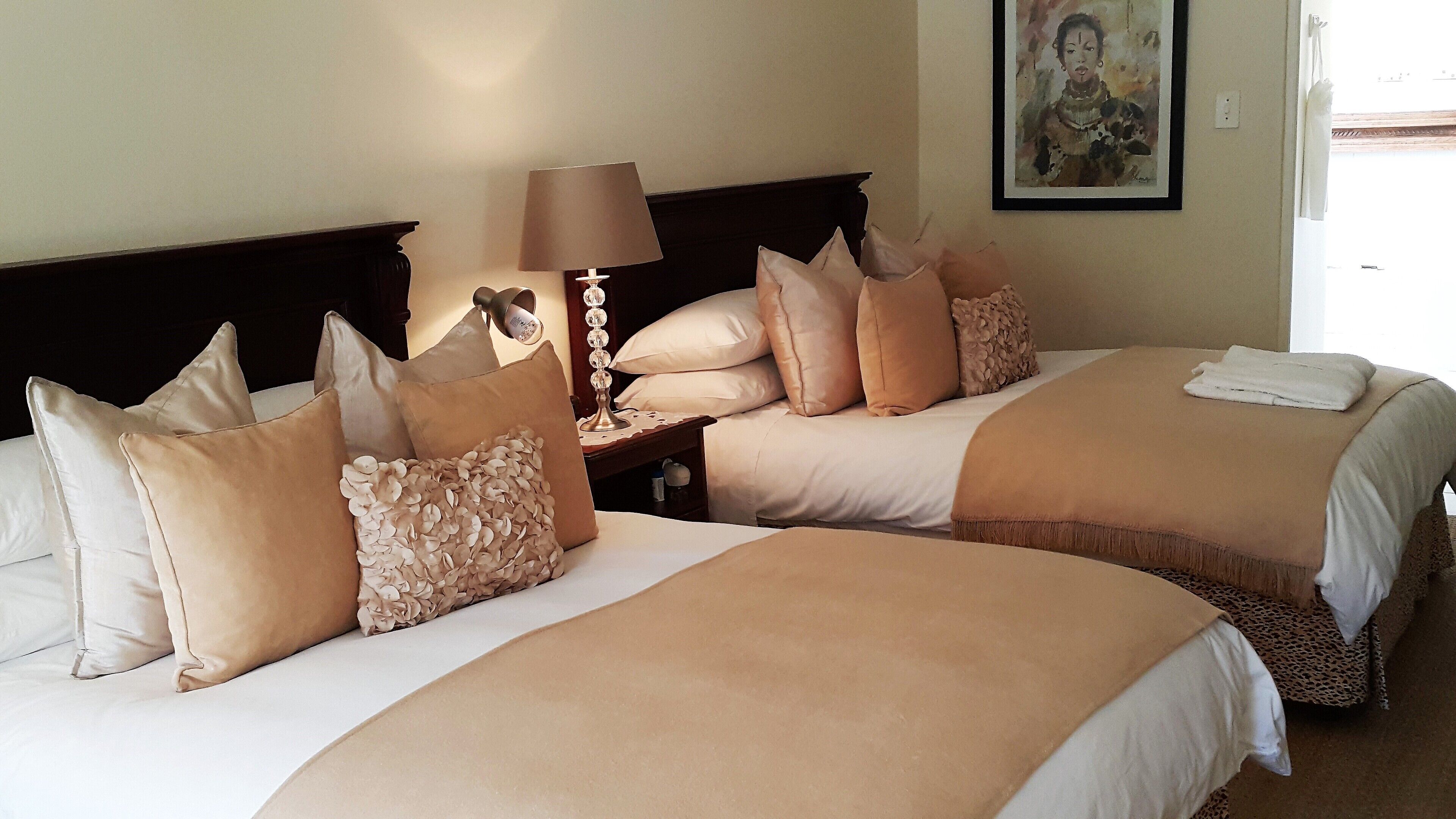 Dos camas con ropa de cama color beige y almohadas decorativas, una mesita de noche con lámpara y un cuadro en la pared.