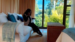 A room with a white sofa, a wicker side table, and a view of a lush green landscape through large windows. - Riversong Guest House (Cape Town)