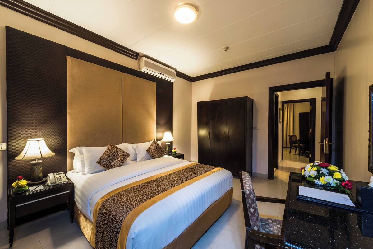 A hotel room with a large bed, two bedside tables with lamps, a dark wooden wardrobe, a desk with a mirror, and a flower arrangement.