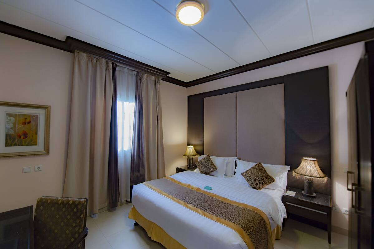 A hotel room with a large bed, a nightstand with a lamp, a chair, and a painting on the wall.