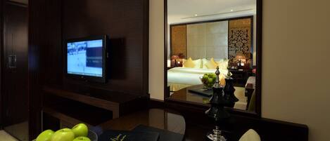 A hotel room with a TV, a bed with white linens, a mirror, and a table with a bowl of green apples and a book.