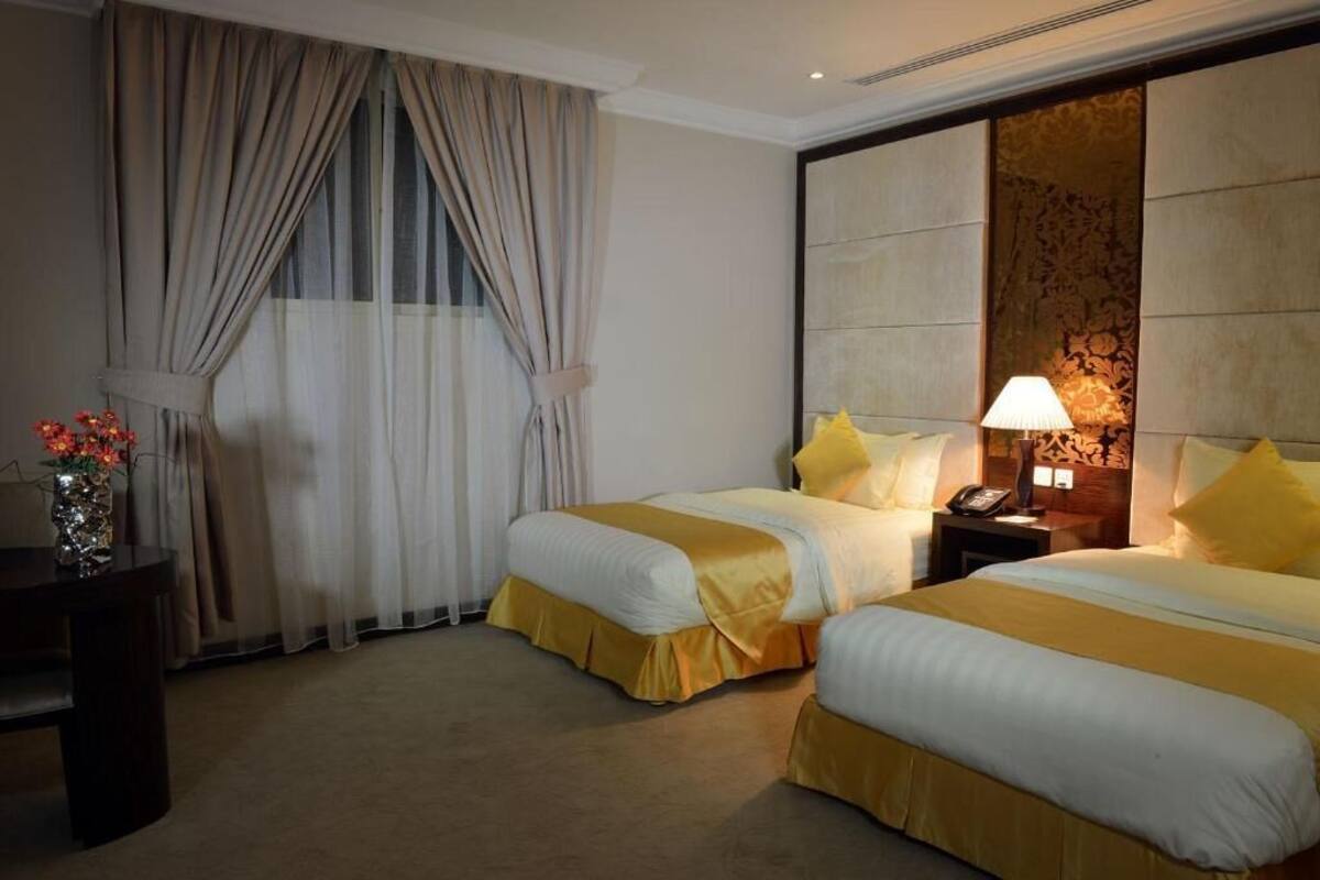 A hotel room with two beds, a nightstand with a lamp, a vase with flowers, and a large window with curtains.
