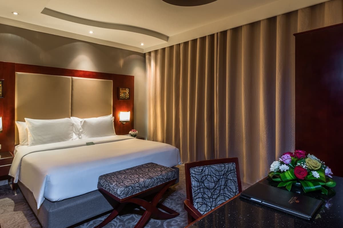 A hotel room with a large bed, bedside lamps, a desk with a laptop, a chair, and a bouquet of flowers.